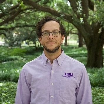 Man in purple shirt smiling and standing in front of a tree and bushes